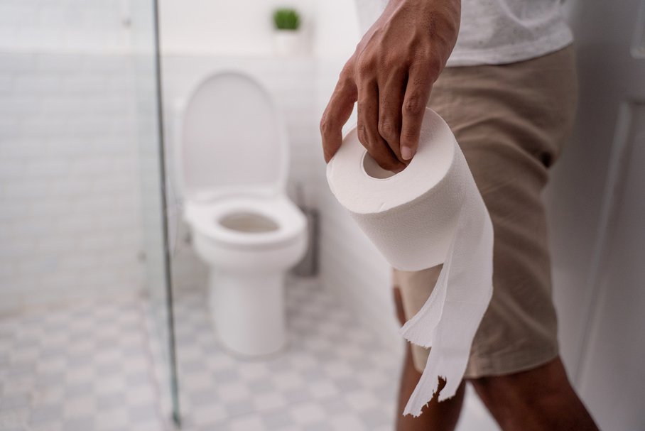 Mężczyzna korzystający z toalety i trzymający w dłoni papier toaletowy.