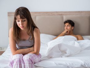 Smutna młoda kobieta siedząca na łóżku w którym leży zamyślony mężczyzna.